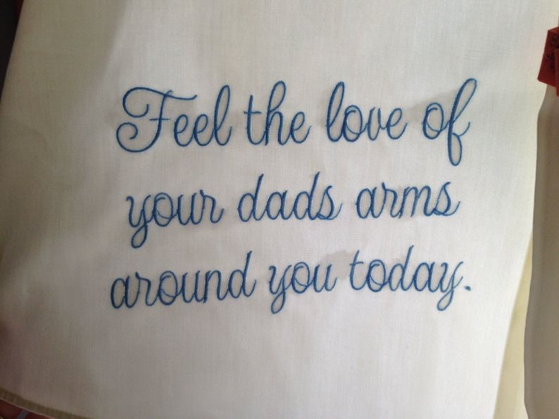 Puede incluir: Tela blanca con texto bordado en azul que dice "Siente el amor de los brazos de tu pap&aacute; alrededor tuyo hoy."