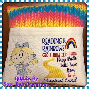 Pode incluir: Uma almofada branca com um bolso na frente. A almofada é decorada com uma imagem bordada de uma menina com tranças, um arco-íris e um caminho de tijolos amarelos. O texto "READING & RAINBOWS Go Hand In Hand They Both Will Take You To A Magical Land" é bordado na almofada. A almofada é feita por Sew Deb Creations.