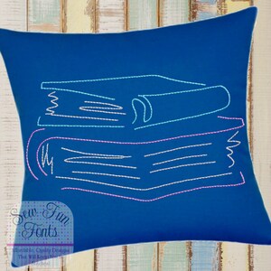 Puede incluir: Una almohada azul con un diseño bordado en blanco y rosa de una pila de libros. El texto "Sew Fun Fonts" está bordado en la esquina inferior izquierda de la almohada.