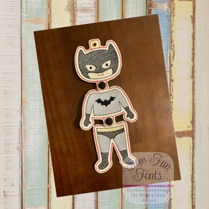 May include: A gray and black felt applique of a superhero character with a bat symbol on the chest. The applique is designed to be used for a craft project.