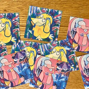 Op de afbeelding: Negen kleurrijke stickers met cartoonfiguren met verschillende uitdrukkingen. De stickers bevinden zich op een houten oppervlak. De figuren zijn geel en roze met blauwe, paarse en groene accenten.