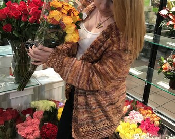 May include: A woman in a brown and orange striped cardigan is holding a bouquet of yellow and orange roses. She is standing in a flower shop and there are other bouquets of flowers in the background.