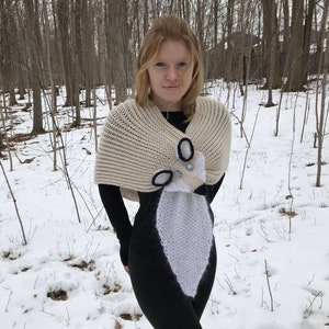 May include: A white knitted shawl with a white and black knitted animal design. The animal has black eyes and a silver button nose.