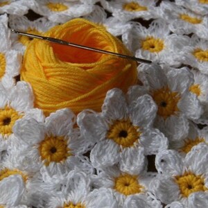 May include: A close-up of a crocheted white daisy flower pattern with yellow centers. A ball of yellow yarn and a crochet hook are in the center of the image.