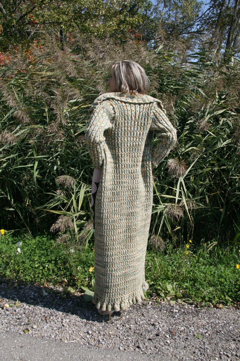 May include: A long, green and brown knitted cardigan with a ruffled hem. The cardigan is worn by a person standing in a grassy area.