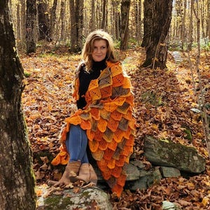 May include: A woman wearing a black shirt and blue jeans sits on a rock in a forest. She is wrapped in a crocheted blanket with orange, brown, and beige squares.