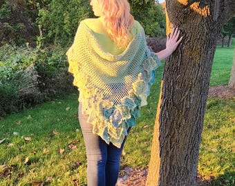 May include: A woman wearing a crocheted shawl with a green, blue, and yellow color gradient. The shawl has a scalloped edge and is draped over her shoulders. She is standing in a grassy area with a tree behind her.