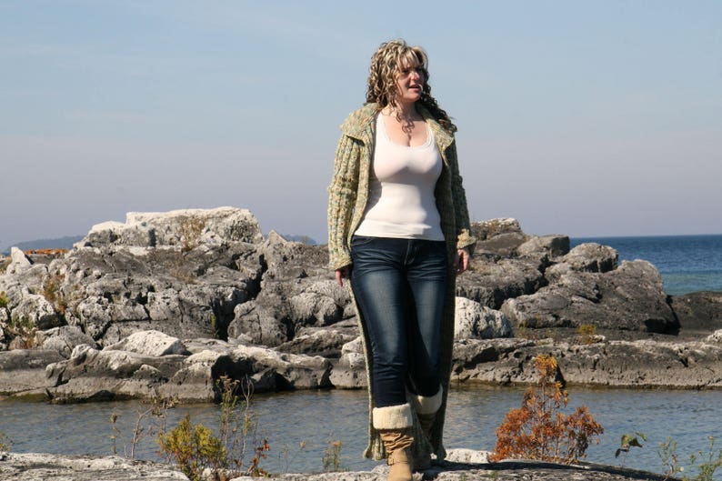 May include: A woman wearing a light green cardigan over a white tank top and blue jeans. She is standing on a rocky shoreline with water in the background.