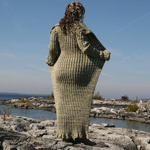 May include: A long, loose-fitting, knitted cardigan in a green and brown color scheme. The cardigan is made of a soft, textured yarn and has a relaxed fit. The cardigan is shown on a person standing on a rocky shoreline.
