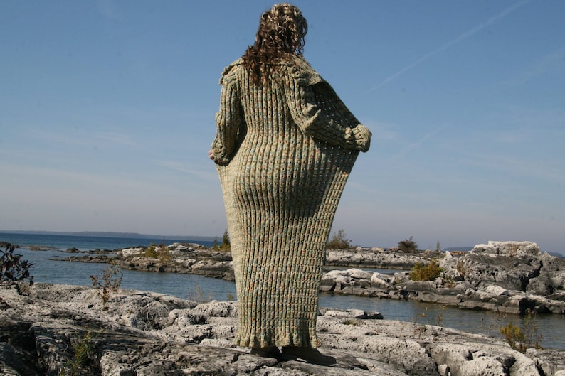 May include: A long, loose-fitting, knitted cardigan in a green and brown color scheme. The cardigan is made of a soft, textured yarn and has a relaxed fit. The cardigan is shown on a person standing on a rocky shoreline.