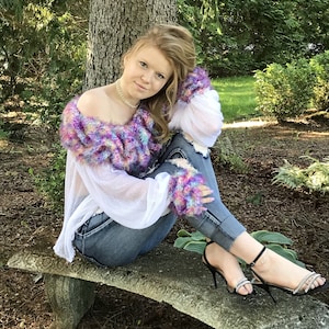 May include: A young person wearing a colourful, fuzzy, off-the-shoulder sweater with a white crocheted overlay, blue jeans, and black heeled sandals with rhinestones.