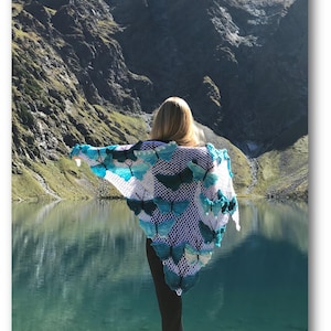 May include: A white crocheted shawl featuring numerous three-dimensional butterfly motifs in various shades of blue, teal, and white. The shawl has an open, net-like stitch pattern and is worn over black leggings and white sneakers.