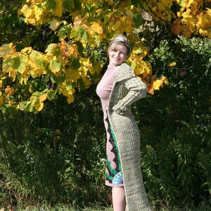 May include: A woman wearing a long, multi-colored cardigan with a pink dress underneath. She is standing in front of a tree with yellow leaves.