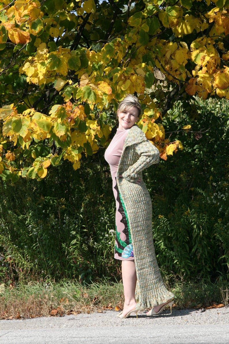 May include: A woman wearing a long, multi-colored cardigan with a pink dress underneath. She is standing in front of a tree with yellow leaves.
