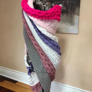 Könnte beinhalten: Ein mehrfarbiger Strick Schal mit einem Farbschema aus Rosa, Weiß, Lila, Grau und Rosa. Der Schal ist über eine schwarze Schaufensterpuppe gelegt.