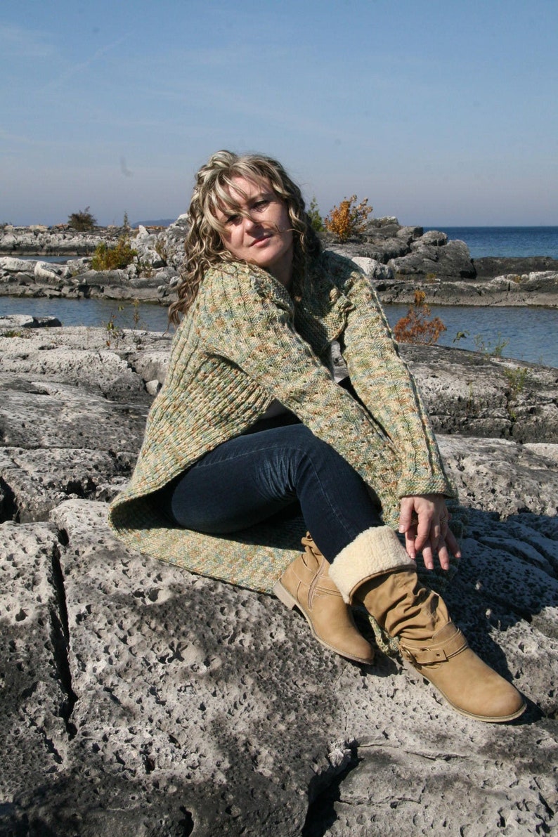 May include: A woman wearing a green and brown patterned sweater with blue jeans and brown boots sits on a rocky shore.