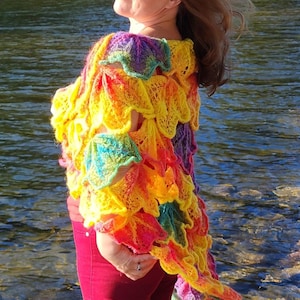 Peut inclure: Un châle crocheté à la main aux couleurs vives : jaune, orange, rose, violet et vert. Le châle présente un motif texturé unique et est porté sur les épaules. La personne porte un pantalon rouge.