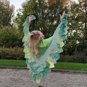 May include: A woman wearing a green and blue crocheted shawl with a scalloped edge, sitting on a swing in a park.
