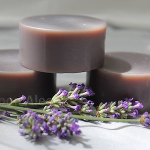 May include: Three round bars of lavender-scented soap stacked on top of each other. The soap is a light purple color and has a smooth, polished finish. The soap is surrounded by lavender flowers.