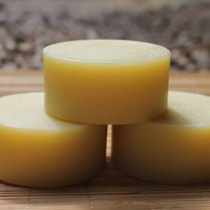May include: Three cylindrical, pale yellow soap bars are stacked on a bamboo mat. The bars have a smooth, matte finish and a uniform shape. The background is blurred, suggesting a natural setting.