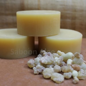 May include: Three round bars of pale yellow soap stacked on top of each other. A pile of small, light brown and white resin pieces are in front of the soap bars. The text "Saboon Aloe" is visible on the bottom soap bar.