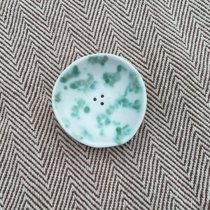 Puede incluir: Un pequeño jabonera de cerámica blanca con un esmalte moteado verde y marrón. La jabonera tiene tres pequeños agujeros en la parte inferior para el drenaje.