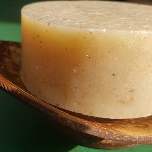 May include: A round, off-white bar of soap with a grainy texture sits on a wooden dish. The soap is in a natural, rustic setting.