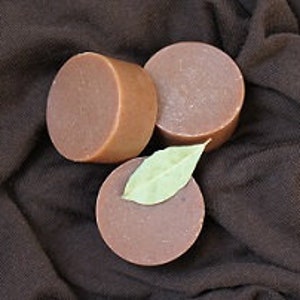 May include: Three round brown bars of soap, two are on a brown fabric surface and one is on top of a green leaf.