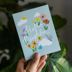 May include: A light blue card with a floral design and the text "Hello Neighbor!" in white cursive. The card features a sun, clouds, flowers, and a butterfly.