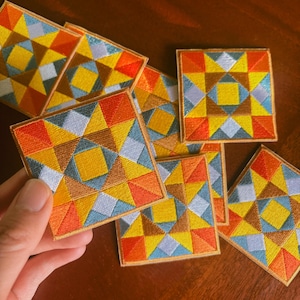 May include: A collection of square embroidered patches with a geometric design. The patches feature a colorful pattern of triangles in shades of yellow, orange, blue, and brown, with a gold border. The patches are arranged on a wooden surface.