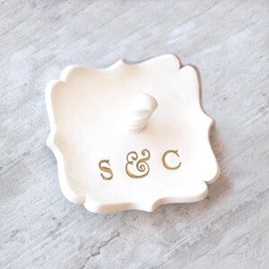 May include: A white ceramic ring holder with a scalloped edge and a central post. The holder features gold lettering with the initials "S & C". The ring holder is a decorative item for storing rings and small jewelry.