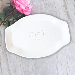 May include: A white oval ceramic tray featuring the embossed letters C & J and the date 10.15.20 centered on the surface. The tray has a smooth finish and slightly raised edges, accompanied by a single pink rose with green leaves.