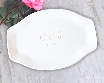 May include: A white oval ceramic tray featuring the embossed letters C & J and the date 10.15.20 centered on the surface. The tray has a smooth finish and slightly raised edges, accompanied by a single pink rose with green leaves.