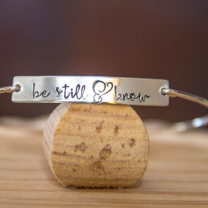 Può includere: Braccialetto rigido in argento con un ciondolo a barra in argento che recita "be still & know".