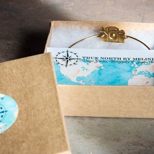 May include: A gold-colored camera charm bracelet is displayed inside a small, open cardboard box. The box contains a card with a map design and the text "TRUE NORTH BY MELINDA Unique Jewelry, Photography & Graphic Art."