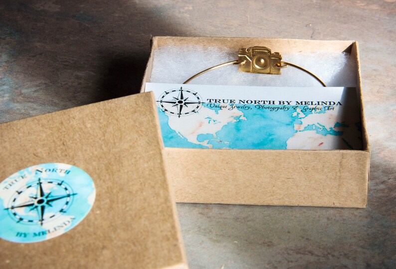 May include: A gold-colored camera charm bracelet is displayed inside a small, open cardboard box. The box contains a card with a map design and the text "TRUE NORTH BY MELINDA Unique Jewelry, Photography & Graphic Art."