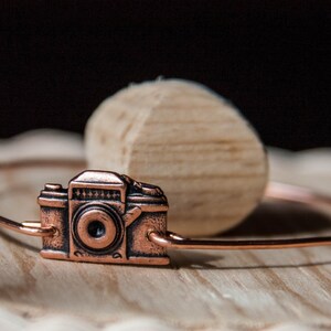 May include: A copper-colored bracelet featuring a miniature camera charm. The camera charm has detailed features and is attached to a thin, circular band. The bracelet rests on a wooden surface with a light-colored, rounded wooden object in the background.