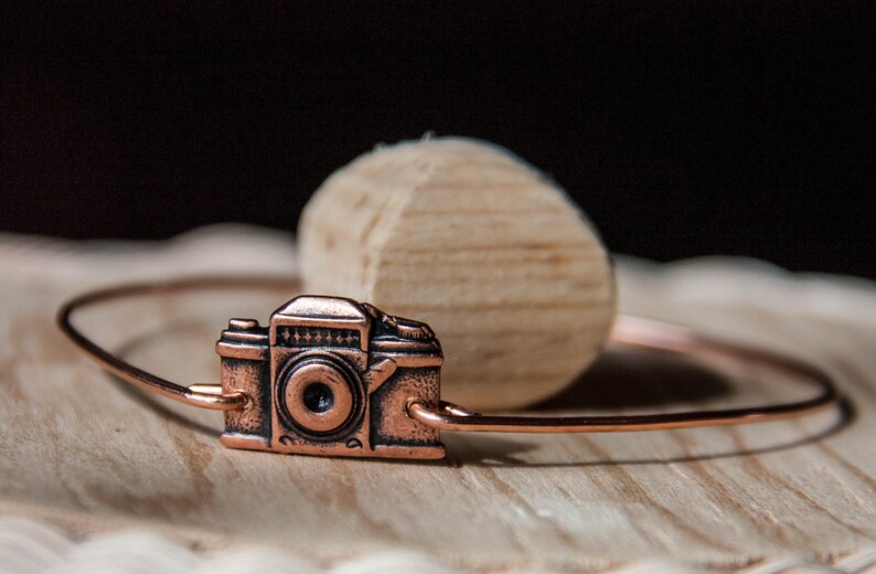 May include: A copper-colored bracelet featuring a miniature camera charm. The camera charm has detailed features and is attached to a thin, circular band. The bracelet rests on a wooden surface with a light-colored, rounded wooden object in the background.