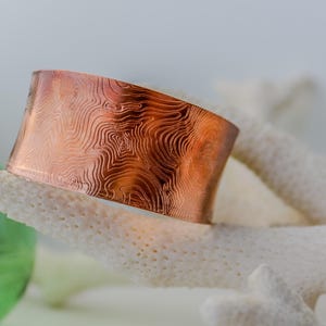May include: A copper cuff bracelet with a swirling, textured pattern. The bracelet is a warm, reddish-brown color and has a smooth, polished finish.