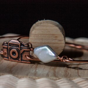 May include: A copper-colored bracelet featuring a detailed camera charm and a large, irregular white pearl. The bracelet is crafted from thin, coiled wire. The camera charm has intricate details, and the pearl adds a touch of elegance.
