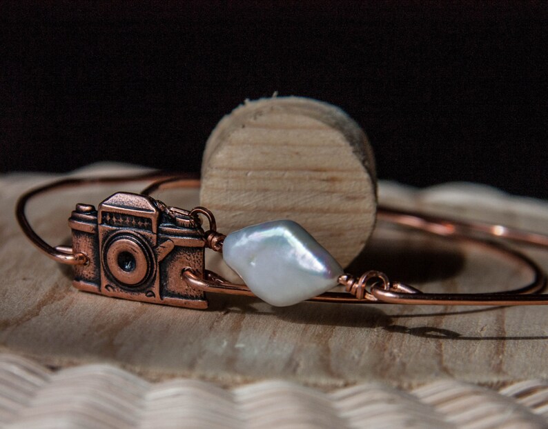 May include: A copper-colored bracelet featuring a detailed camera charm and a large, irregular white pearl. The bracelet is crafted from thin, coiled wire. The camera charm has intricate details, and the pearl adds a touch of elegance.