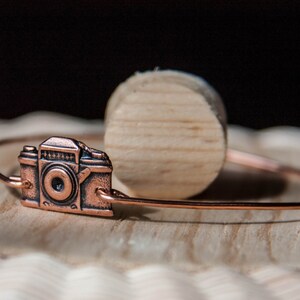 May include: A copper-colored bracelet featuring a detailed camera charm. The charm is a miniature replica of a vintage camera. The bracelet is displayed on a wooden surface, with a wooden cylinder in the background.
