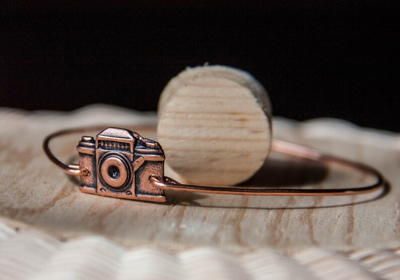 May include: A copper-colored bracelet featuring a detailed camera charm. The charm is a miniature replica of a vintage camera. The bracelet is displayed on a wooden surface, with a wooden cylinder in the background.