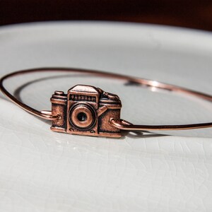 May include: A copper-colored bracelet featuring a vintage camera charm. The camera charm is detailed with a lens and buttons, and the bracelet is a simple, thin band. The jewelry is displayed on a white, textured surface.