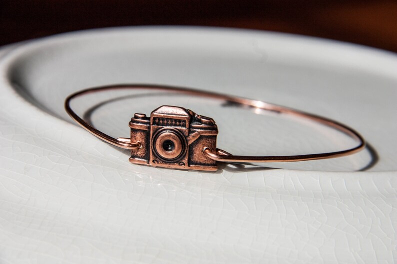 May include: A copper-colored bracelet featuring a vintage camera charm. The camera charm is detailed with a lens and buttons, and the bracelet is a simple, thin band. The jewelry is displayed on a white, textured surface.