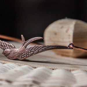 May include: A copper-toned bracelet with a detailed, stylized bird design. The bracelet is open and has a simple, curved shape.