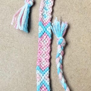 May include: Handmade friendship bracelet in shades of pink, blue, and white. The bracelet features a woven design with a braided loop and tassel ends. A colorful accessory for everyday wear.