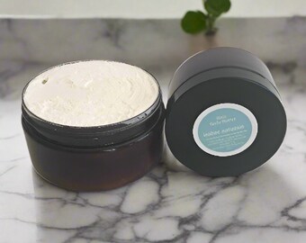 May include: A black jar with a white label that reads "Body Butter, Luxe Naturals" filled with a white cream. The jar is sitting on a white marble surface.