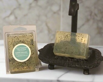 May include: A clear bar of soap with black poppy seeds in a clear plastic wrapper. The soap is labeled "Citrus Poppyseed Glycerin Soap with Olive Oil" and "leabee naturals". The soap is sitting on a black soap dish with a red faucet handle.