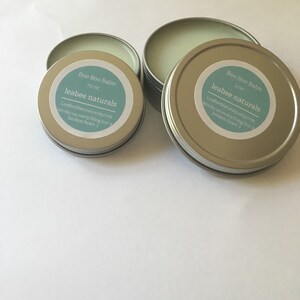 May include: Several small, round, silver tins of "Boo Boo Balm" are displayed. Each tin has a light blue label with the product name, size, and the "leabee naturals" brand name. The text on the label also includes a website address and a tagline.